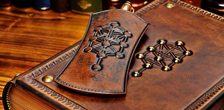 Thick leather book and pen holder lay down to the table with books in the background. The tree of life symbol are on both items.